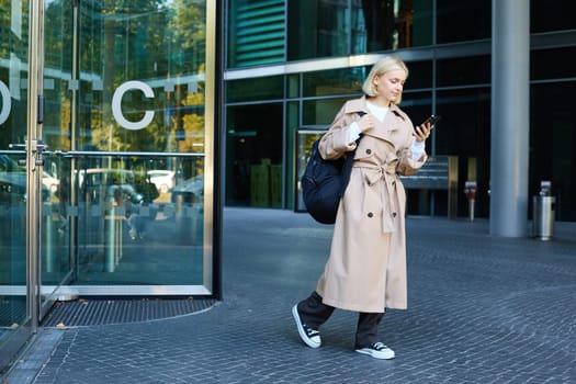 Portrait of blond young woman walking on street, student with backpack looking at her mobile phone, reading message on smartphone.