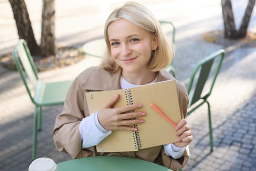 Outdoor shot of young beautiful blond girl with notebook, woman sitting in outdoor cafe, writing in journal, making plans in her planner, putting notes.