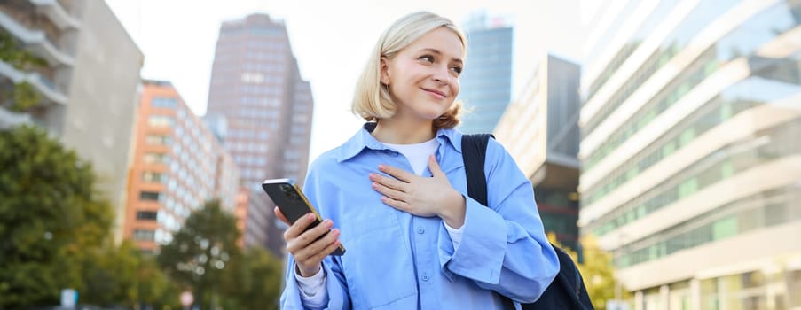 Image of young smiling woman, young professional, standing with backpack and mobile phone, holding hand on chest, looking aside with pleased, happy face expression.