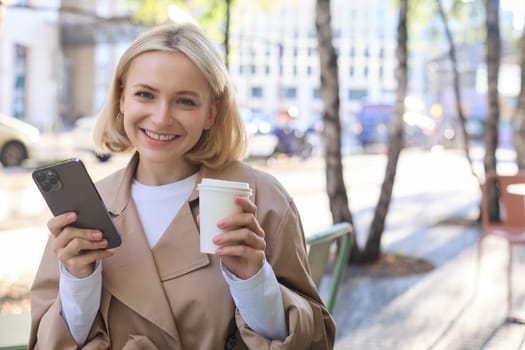 Portrait of cute blond woman with smartphone, drinking chai, coffee at favourite place in city centre, sitting outdoors, holding mobile phone.