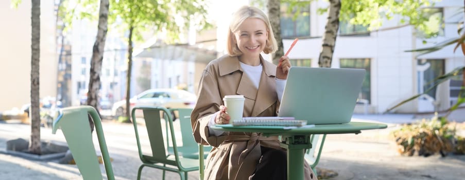 Portrait of young smiling blond woman, working on laptop, sitting in outdoor cafe on street, drinking coffee. Lifestyle and people concept