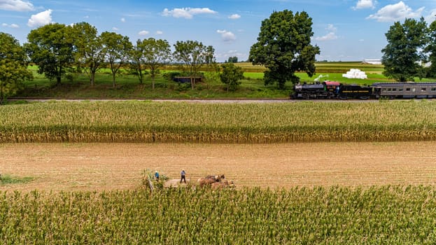 Ronks, Pennsylvania, September 11, 2021 - An Aerial View of Six Horses Pulling an Amish Harvesting Corn Stalk Machine, as Steam Passenger Train Travels
