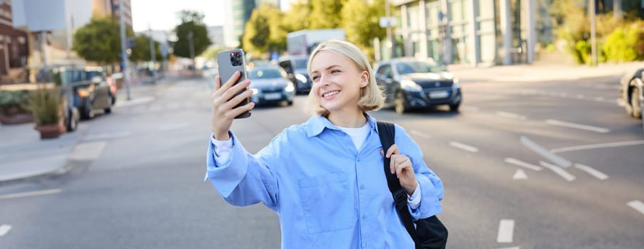 Image of smiling young woman, modern girl with smartphone, taking selfie, shooting a picture of busy city streets, recording video outdoors.