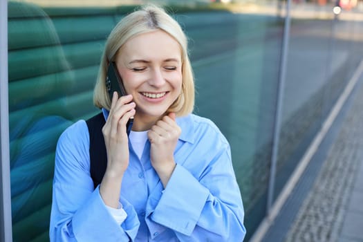 Lifestyle portrait of happy blond woman, chatting over the phone, answers a call, laughing and smiling, standing on street and looking cheerful.