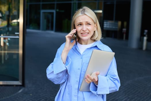 Image of young smiling woman, working in company, standing near office with laptop, answers phone call, chatting with someone.
