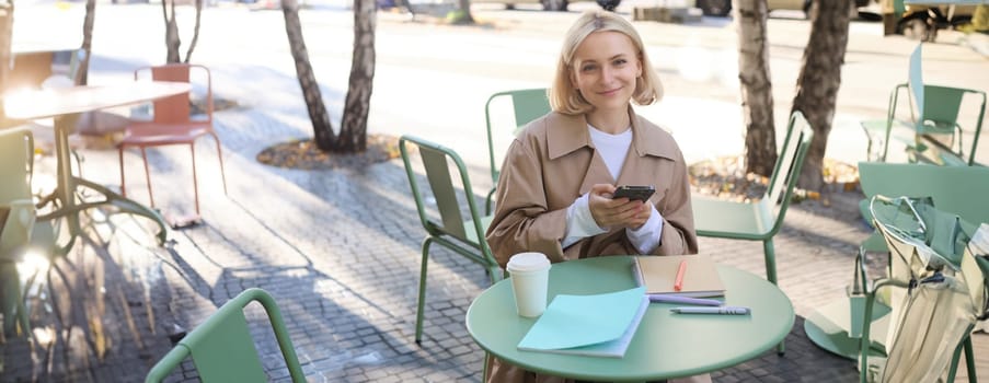 Portrait of smiling young woman using mobile phone while doing homework, working on project, sitting outdoors in cafe, drinking coffee and studying, holding smartphone.