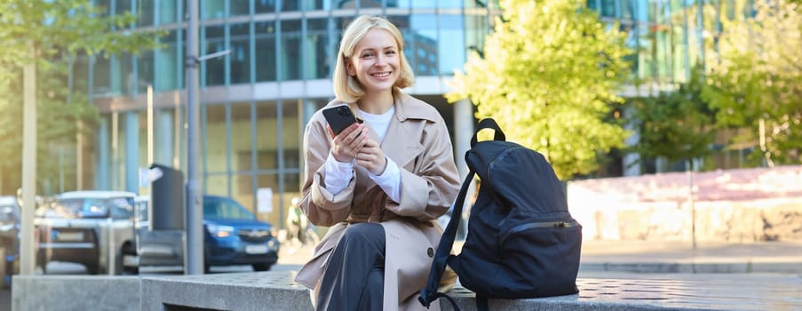 Outdoor shot of young blonde woman on street, sitting on bench with mobile phone, smiling at camera, waiting for friend outside of building, having a break.