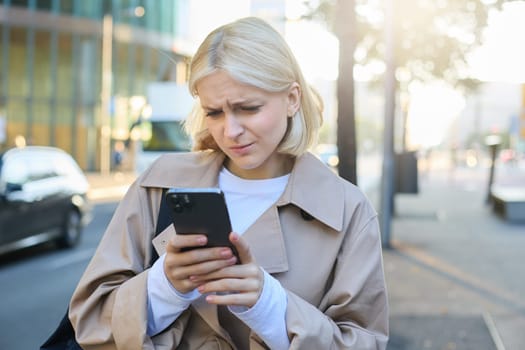 Portrait of young female model on street, looking at smartphone with disappointed, upset face, frowning while reading mobile phone message.