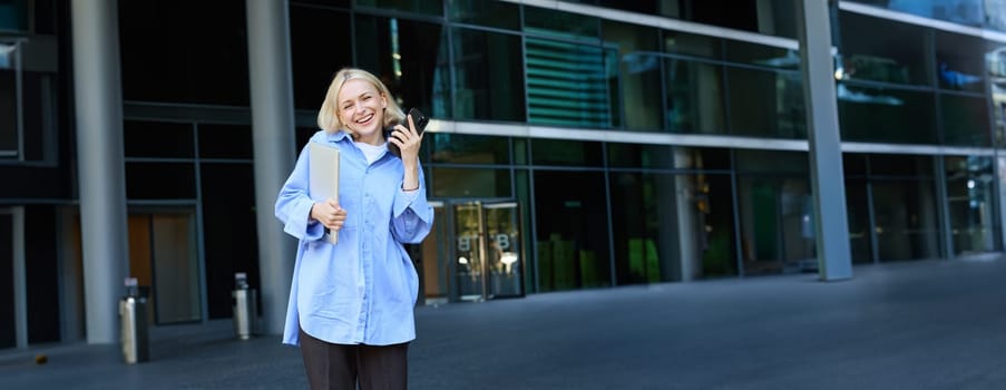 Vertical shot of modern, stylish young blond woman smiling, posing in trousers and blue collar shirt, holding smartphone and laughing, posing near campus building, has laptop in hand.
