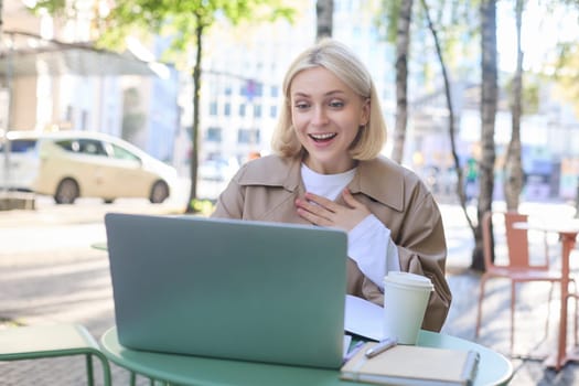 Young woman, freelancer, student doing homework in outdoor cafe, drinking her coffee on street, using laptop, connects to online lecture or doing course in internet.