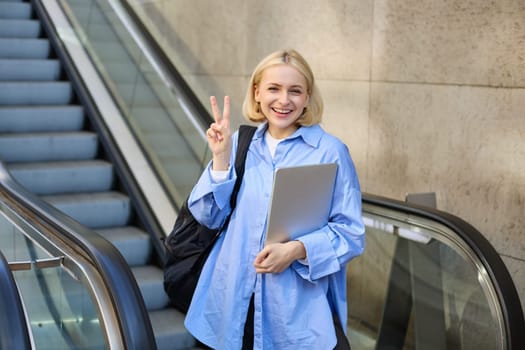Beautiful young blond woman in blue collar shirt, going to work, using escalator, standing with laptop and showing peace sign.