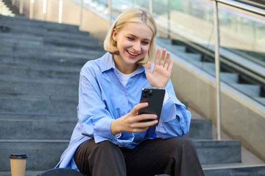 Cute young woman, looking at smartphone, waving at mobile camera, saying hello, online video chatting, smiling happily.
