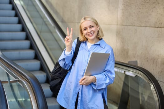 Lifestyle portrait of positive young woman, female student with laptop and backpack, showing peace sign, standing near escalator.