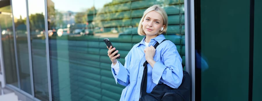 Portrait of beautiful blond girl, smiling woman in wireless earphones, listening to music on smartphone, waiting for friend on street, standing outdoors, leaning on building.