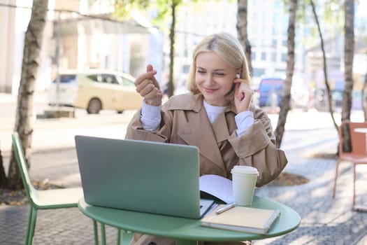 Cheerful young woman in trench coat with coffee, using wireless headphones and laptop, working remotely, connect to online class, video chatting.