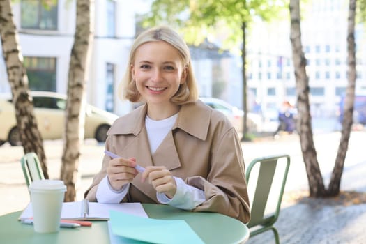 Image of young blonde woman in stylish trench, sitting in outdoor cafe, drinking coffee and working on documents, studying, doing homework in her notebook, smiling at camera.
