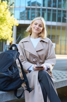 Vertical shot of young blond woman sitting on street bench with mobile phone, has backpack, waiting for someone, order taxi in smartphone application and looking around, smiling carefree.