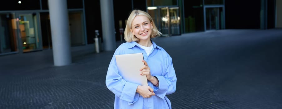 Portrait of young blond woman, student standing near her campus with notebooks and documents, wearing blue shirt and smiling at camera. Education concept