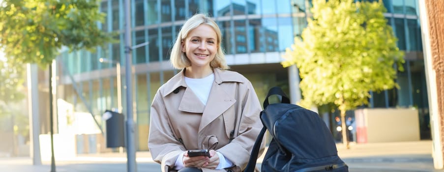 Lifestyle portrait of smiling female model sitting on street bench with backpack and mobile phone, waiting for friend and scrolling social media on smartphone.