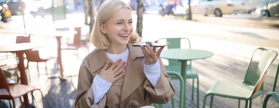 Lifestyle portrait of happy young woman, sitting in cafe and recording voice message, holding mobile phone near lips and talking into microphone, spending time in cafe, drinking coffee and chatting.