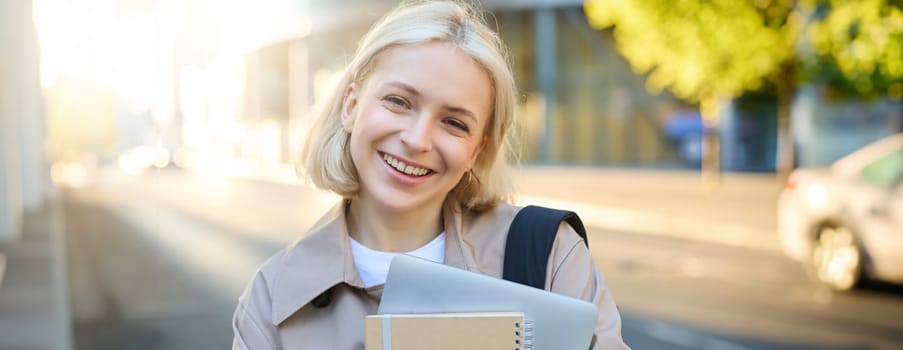 Close up portrait of smiling blond woman, standing on street with notebooks, carries journal and work documents, looking happy at camera.