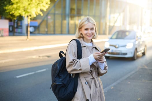 Stylish young blonde woman, girl with smartphone and backpack, standing on street, using mobile phone application, posing close to the road with cars and sky scrappers.
