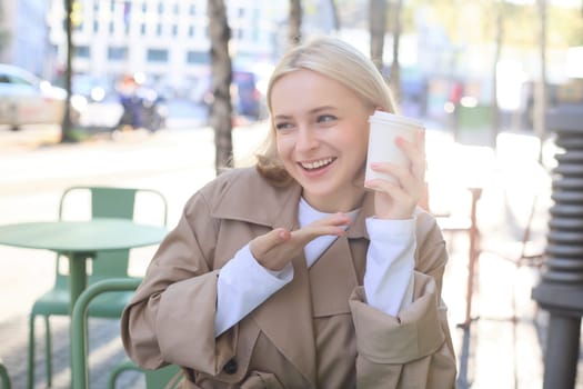 Portrait of smiling blond woman, pointing finger at her cup of coffee, orders takeaway, sits in street cafe, shows her favourite drink.