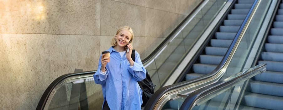 Smiling young woman on escalator, talking on mobile phone, drinking coffee and looking happy, going to university or college, on her way in city.
