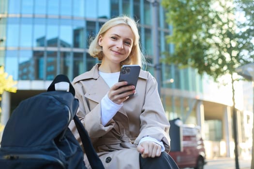 Portrait of smiling young woman, sitting on street bench, holding mobile phone, chatting with someone on smartphone while waiting for friend outside.