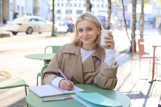 Image of young smiling woman, female entrepreneur writing, making corrections in documents, using notebook, drinking coffee and sitting in outdoor cafe.