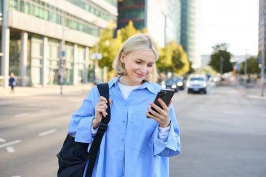 Portrait of young city girl, urban woman with backpack, reading messages on smartphone and smiling, checking notifications, standing on street.