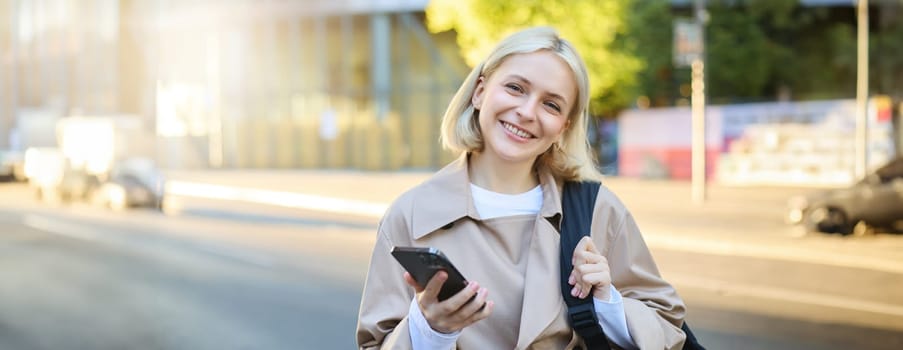 Close up portrait of young smiling college girl, student with backpack, holding smartphone, looking happy at camera, waiting on street.