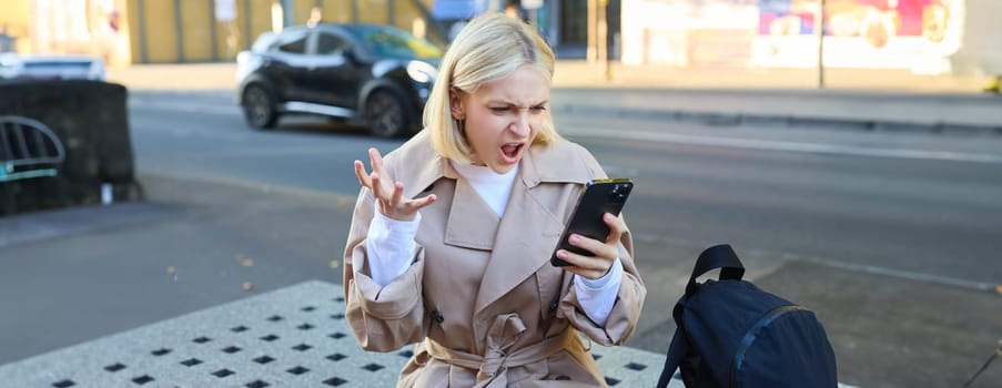 Portrait of angry young woman, shouting at her mobile phone in frustrated, being outraged, sitting on street bench outdoors.