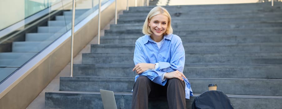 Urban style portrait of young woman, student sitting on city street stairs, with backpack, cup of coffee and laptop, looking happy and upbeat, smiling.