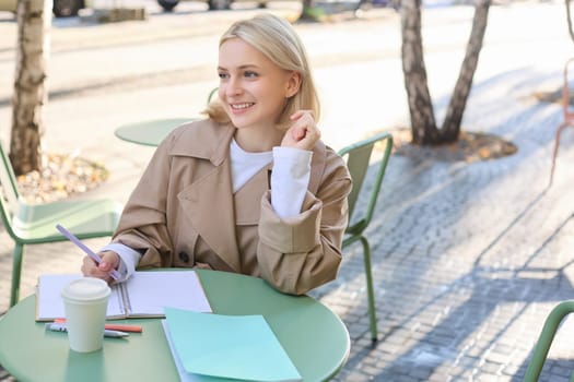 Portrait of carefree, blond cute woman working with documents, drinking takeaway coffee and sitting in cafe outdoors with documents, writing essay, student doing homework.