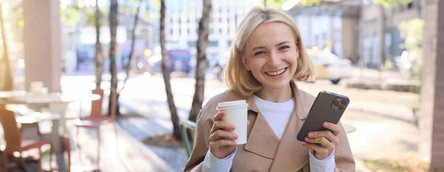 Portrait of blond smiling woman with smartphone, holding cup of coffee, drinking chai and enjoying sunny day outdoors in city centre.