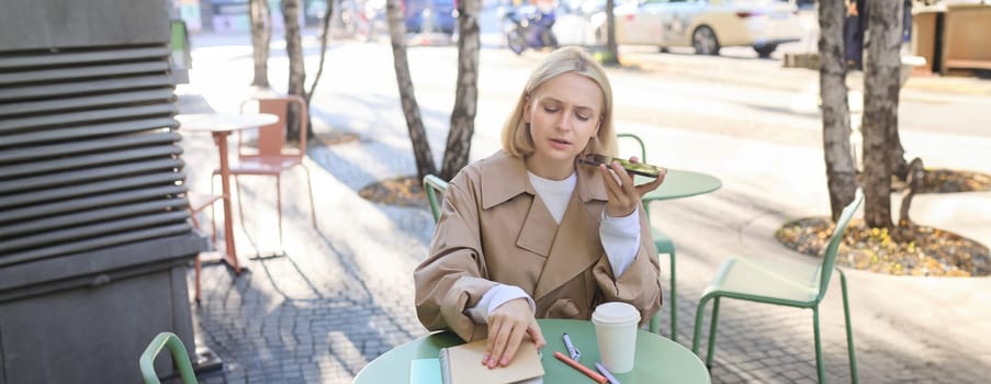 Portrait of serious, concentrated woman, opens her notebook, records voice message, sending notification to coworker, drinking coffee, working outdoors.