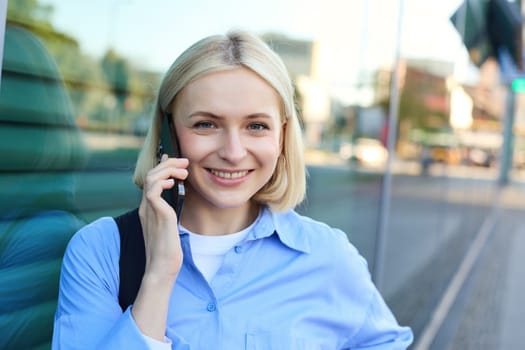 Close up portrait of smiling blond woman, chatting on the phone, talking on mobile telephone, standing on street outdoors.