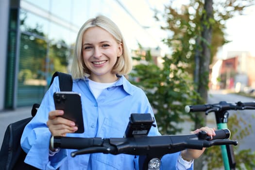 Modern young woman scans QR code with smartphone app to rent an electric scooter on street, smiling and looking happy.