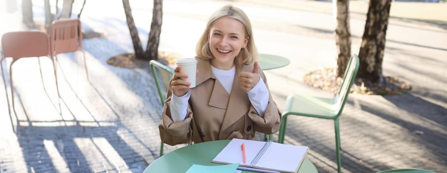 Smiling, cheerful woman drinking her coffee in street cafe, writing in notebook, doing homework, making notes. Student and lifestyle concept.