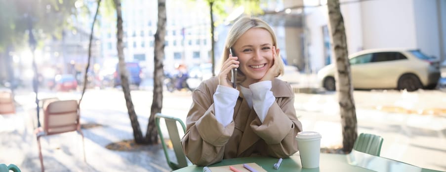 Portrait of young beautiful woman in cafe, sitting outdoors on bright sunny day, talking on smartphone, chatting over the phone and smiling.