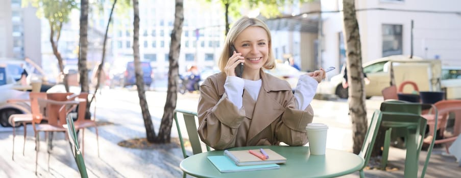 Image of stylish blond woman talking on mobile phone, drinking coffee in cafe outdoors, enjoying warm weather in city centre, answer telephone call on smartphone.