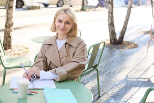 Portrait of young smiling woman, student working, doing homework outdoors, sitting in street cafe, drinking coffee from takeaway cup.