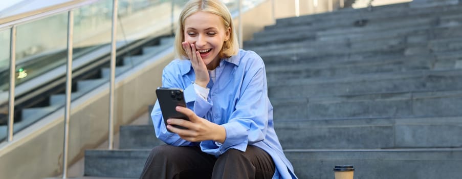 Portrait of young blond woman, sitting outside campus on city stairs, looking at smartphone with happy surprised face, receive positive good news on video chat, watching interesting video.