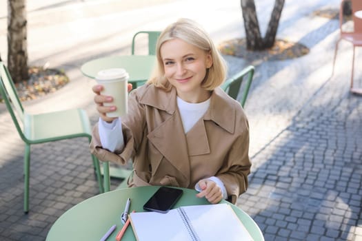 Close up portrait of smiling, beautiful woman raising a cup, showing takeaway coffee and looking happy at camera, drinking favourite beverage in cafe.