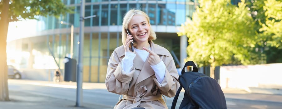 Portrait of cheerful smiling woman, talking on mobile phone, pointing at her smartphone while calling someone, smiling and laughing at camera, sitting on street bench outdoors.