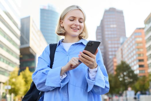 Portrait of beautiful young woman in city centre, standing in blue shirt with smartphone, checking notifications, waiting for car, order taxi on mobile phone app.