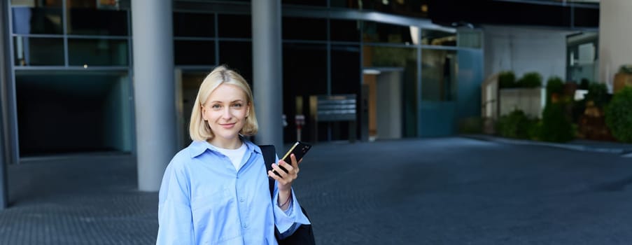 Vertical portrait of young office manager, woman in blue collar shirt and backpack, holding laptop and smartphone, waiting near business building on street, smiling at camera.