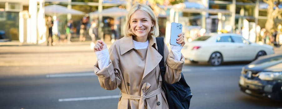 Happy female student on busy street, holding mobile phone, raising hands up and cheering, chanting, winning and celebrating, smiling pleased at camera.