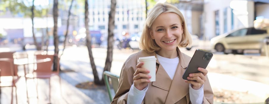 Portrait of cute blond woman with smartphone, drinking chai, coffee at favourite place in city centre, sitting outdoors, holding mobile phone.